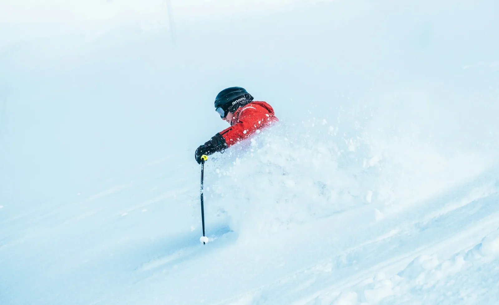 A skier in a red jacket throws up a cloud of fresh powder while turning downhill.