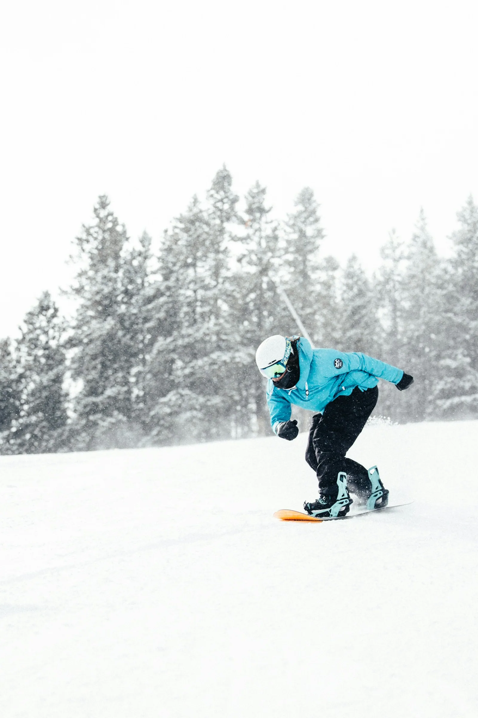 A snowboarder in a turquoise jacket carves through fresh snow in front of evergreen trees.